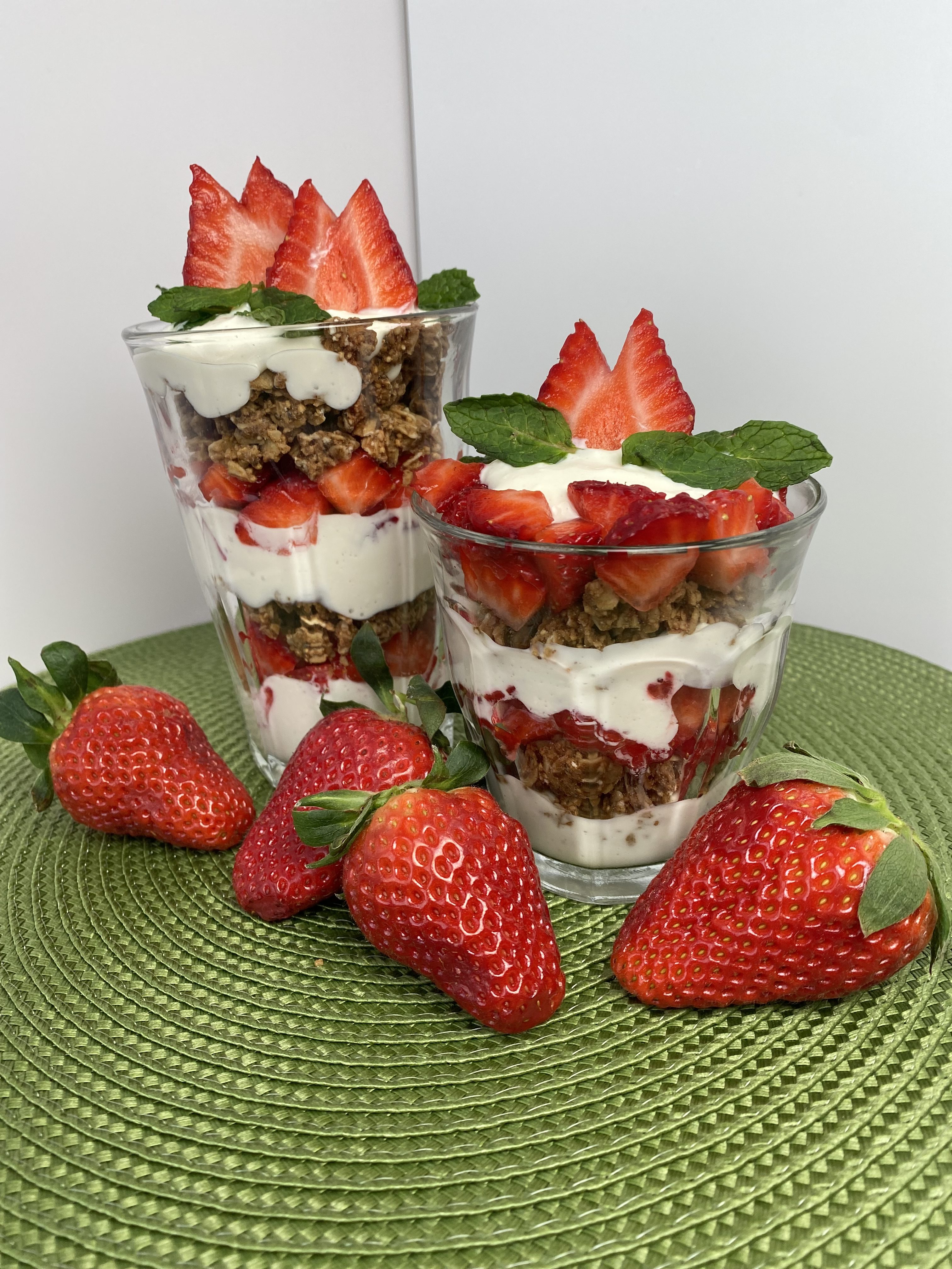 a tall and short glass filled with layered strawberry, yogurt parfait on top of a green mat with whole strawberries in front